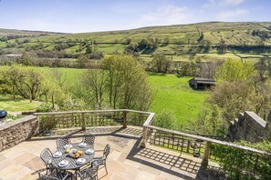 A patio with a table set for dining, overlooking a green landscape with hills and a small building. - 3 Bed in Gunnerside (oc-ds097) (Gunnerside)