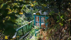 Un pont en bois mène à un petit bâtiment rustique surmonté d’un toit vert, le tout étant entouré d’un feuillage dense.