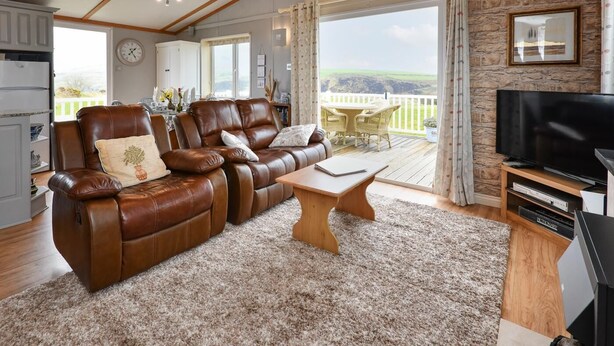Un salon avec un canapé en cuir marron, une table basse en bois et un téléviseur posé sur un meuble en bois. Il y a un balcon avec vue sur la campagne.