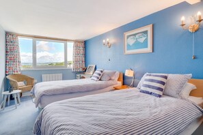 Ein Schlafzimmer mit zwei Betten, einem Korbstuhl, einem kleinen Tisch und einem Fenster mit Vorhängen.