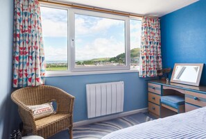 Ein Schlafzimmer mit großem Fenster, einem Korbstuhl, einem Schminktisch und einem Bett.