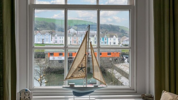 Une maquette de voilier est posée sur un rebord de fenêtre, d’où l’on aperçoit des maisons aux couleurs vives et, au loin, un train.