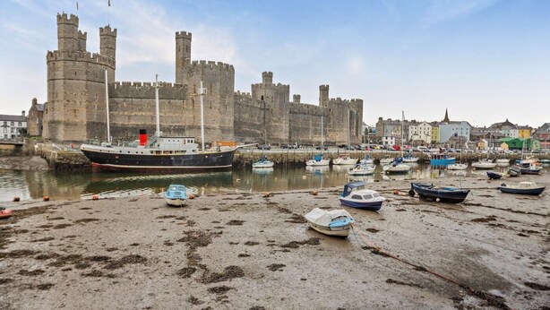 A coastal scene with a large ship docked at a harbor, a castle in the background, and several smaller boats on the shore. - 4 Bed in Caernarfon (oc-30479) (Dinas)