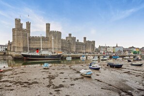 A coastal scene with a large ship docked at a harbor, a castle in the background, and several smaller boats on the shore. - 4 Bed in Caernarfon (oc-30479) (Dinas)