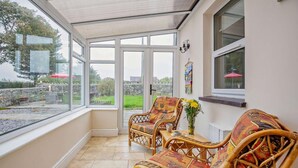 A conservatory with wicker furniture, a table with a vase of flowers, and a view of the garden. - 4 Bed in Caernarfon (oc-30479) (Dinas)