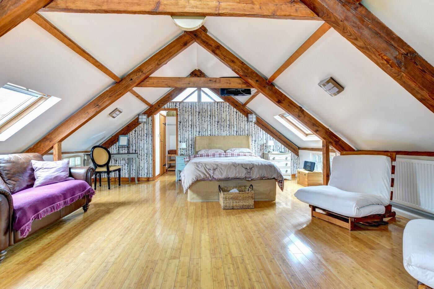 A spacious attic bedroom with a sloped ceiling, wooden beams, a large bed, a desk, and a chair.
