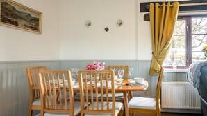 A dining room with a table set for four, wooden chairs, a painting on the wall, and a window with curtains. - 3 Bed in Llanfairpwll (oc-22999) (Star)