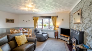 A living room with a fireplace, a sofa, a dining table with chairs, a television, and a window with curtains. - 3 Bed in Llanfairpwll (oc-22999) (Star)