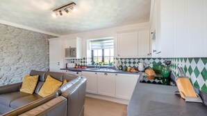 A modern kitchen with white cabinets, a stone wall, a grey sofa with yellow cushions, and a window with a view. - 3 Bed in Llanfairpwll (oc-22999) (Star)
