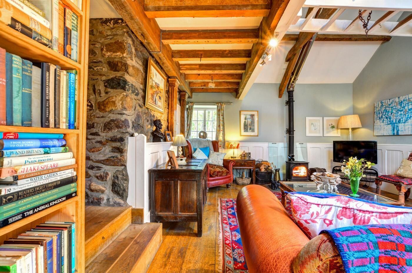 A cozy living room with a stone wall, wooden beams, a fireplace, and a bookshelf filled with books.