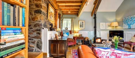 A cozy living room with a stone wall, wooden beams, a fireplace, and a bookshelf filled with books.