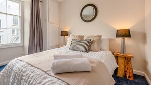 Una cama bien hecha con edredón blanco y almohadas, dos lámparas de noche, un espejo redondo en la pared y una ventana con cortinas.
