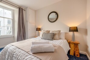 A neatly made bed with a white comforter and pillows, two bedside lamps, a round mirror on the wall, and a window with curtains. - 4 Bed in Tenby (oc-25955) (Tenby)