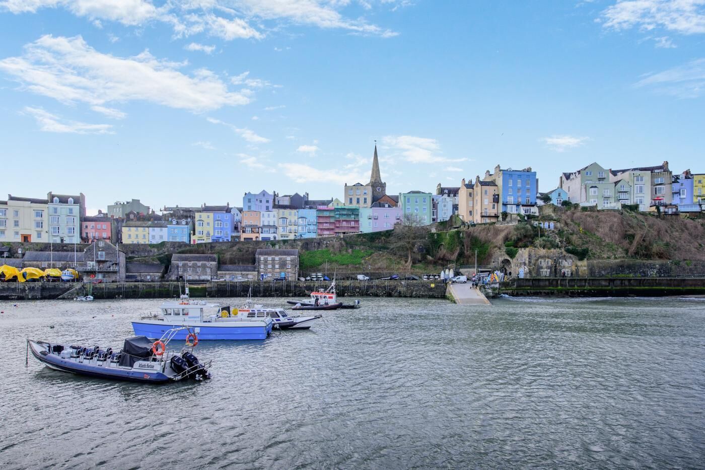 Une ville côtière aux bâtiments aux couleurs vives, avec un clocher d’église et des bateaux sur l’eau.