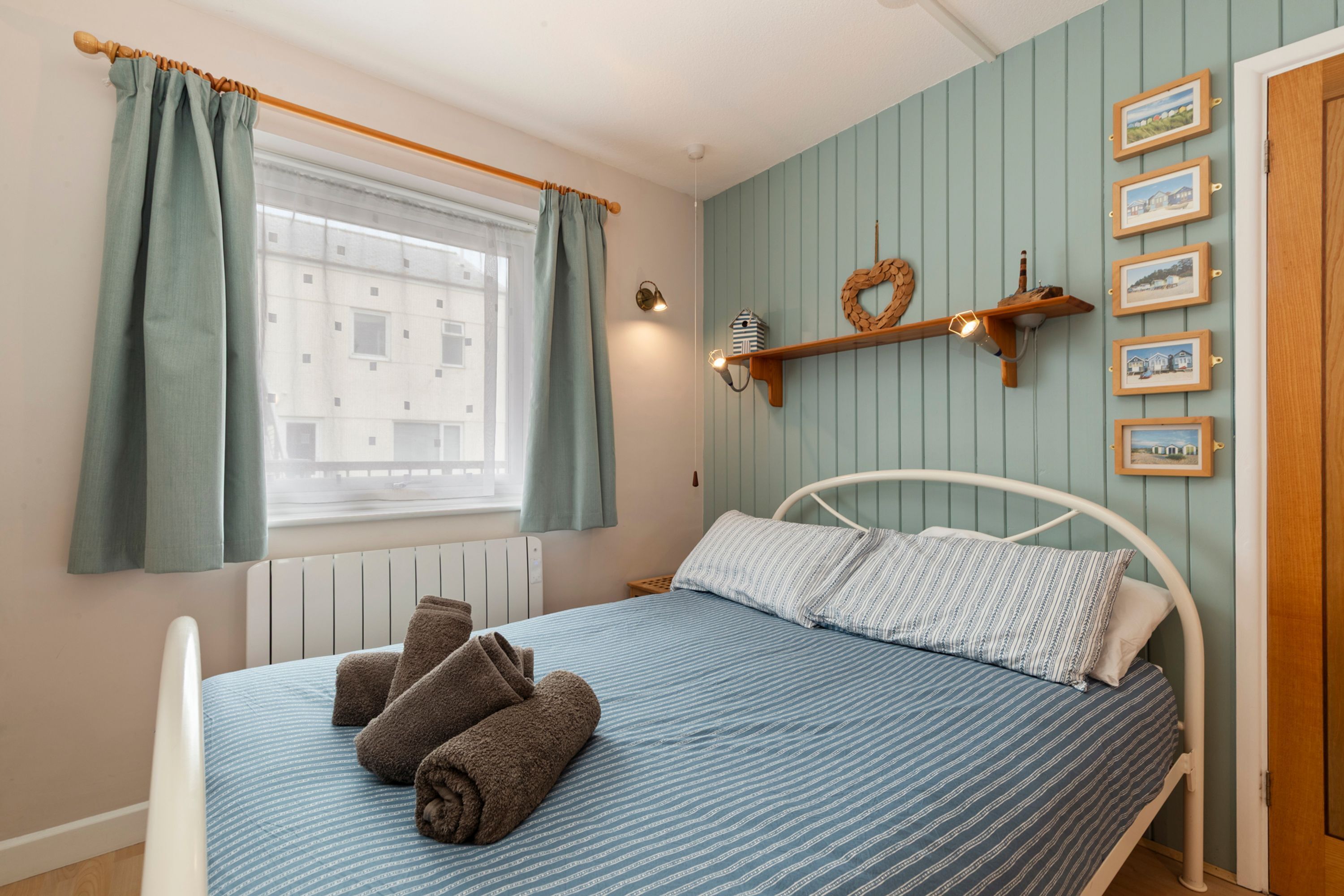 A bedroom with a bed, curtains, a wall-mounted shelf with pictures, and a window.