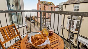 A balcony with a wooden table, a chair, a basket of croissants, a glass of orange juice, and a muffin. - St Mary's Seaview - In Town Walls, Close to Beach (Tenby)