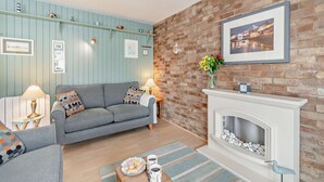 A cozy living room with a blue wall, a brick wall with a fireplace, and a sofa with decorative pillows. - St Mary's Seaview - In Town Walls, Close to Beach (Tenby)