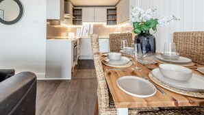 Un comedor con una mesa de madera dispuesta para cuatro, un florero con flores blancas y una cocina al fondo.