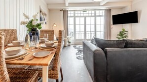 Comedor con mesa de madera dispuesta para cuatro personas, sillas de mimbre, sofá gris y televisor montado en la pared.