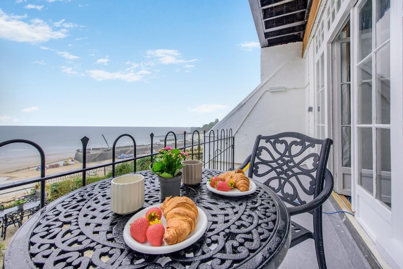 Ein Balkon mit Frühstückstisch, mit Croissants und Erdbeeren, mit Blick auf einen Strand.