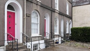 Un edificio con puerta rosa y ventanas de marco blanco, que presenta fachada de piedra y un pequeño jardín.