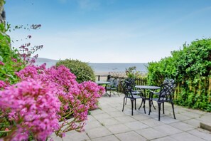 Een terras buiten, met tafels en stoelen, omgeven door groen en bloemen.