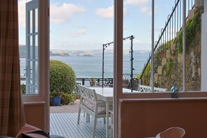 A room with a large window overlooking a coastal area, a small balcony with outdoor seating, and a view of the sea and distant land. - 1 Bed in Tenby (oc-25957) (Tenby)