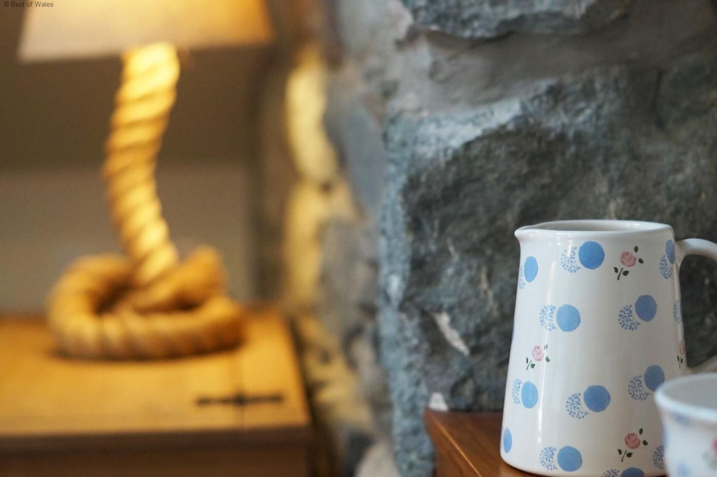A rustic stone wall, a wooden nightstand with a lamp, and a polka-dotted mug.