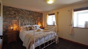 A bedroom with a stone wall, a metal bed frame, a bedside table, a window with curtains, and a nightstand with a lamp. - 9 Bed in Pwllheli (oc-387040) (Pwllheli)