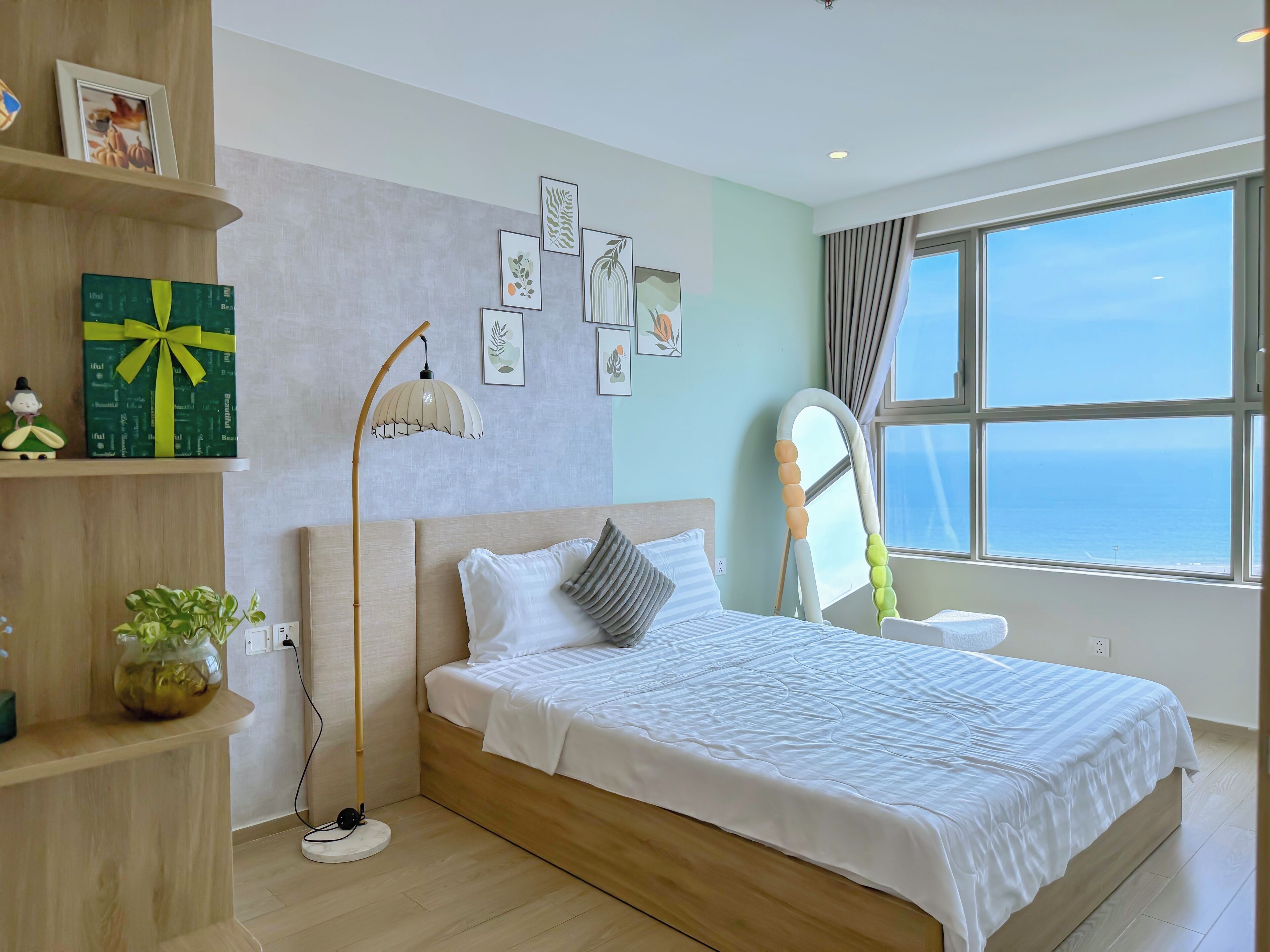 A modern bedroom with a large bed, a bedside lamp, a view of the sea through a window, and a shelf with a gift box.