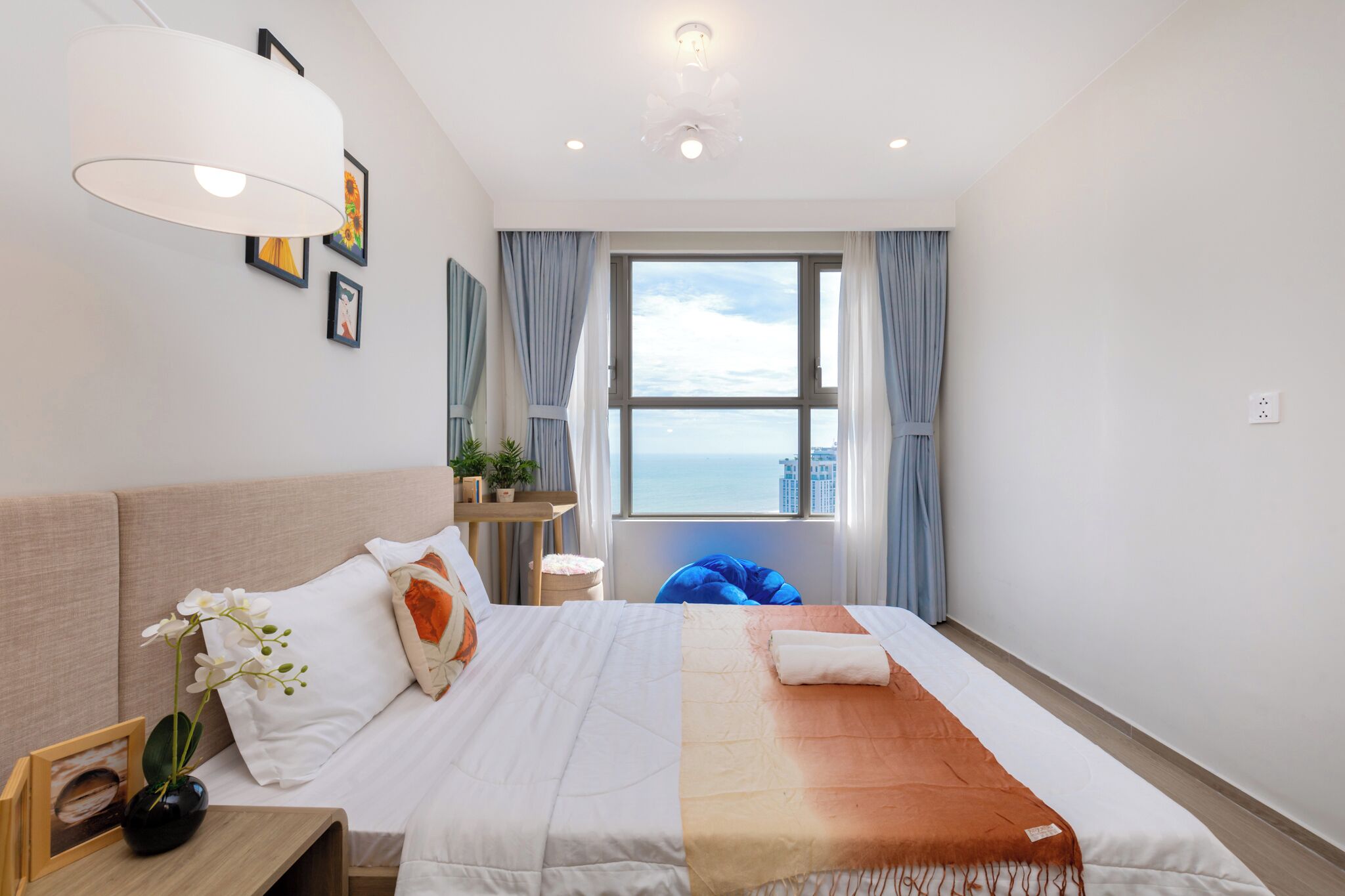 Ein Hotelzimmer mit Bett, Nachttisch mit Blumenvase, Fenster mit Meerblick und Deckenleuchte.