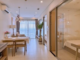 A hotel room with a bed, a desk with a chair, a dining table with chairs, a potted plant, and a view through a glass door.