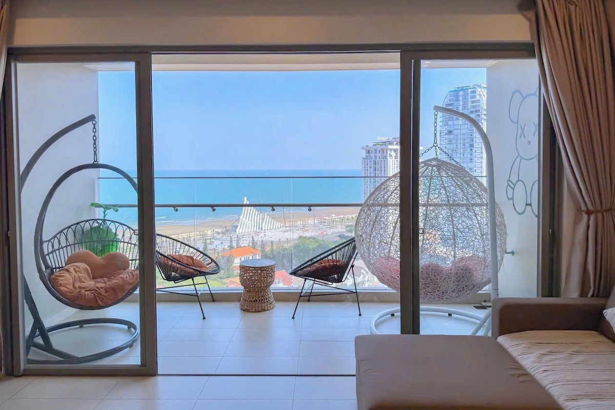 Un balcón con vista al mar y edificios, dos sillas colgantes, una mesita redonda y un sofá.