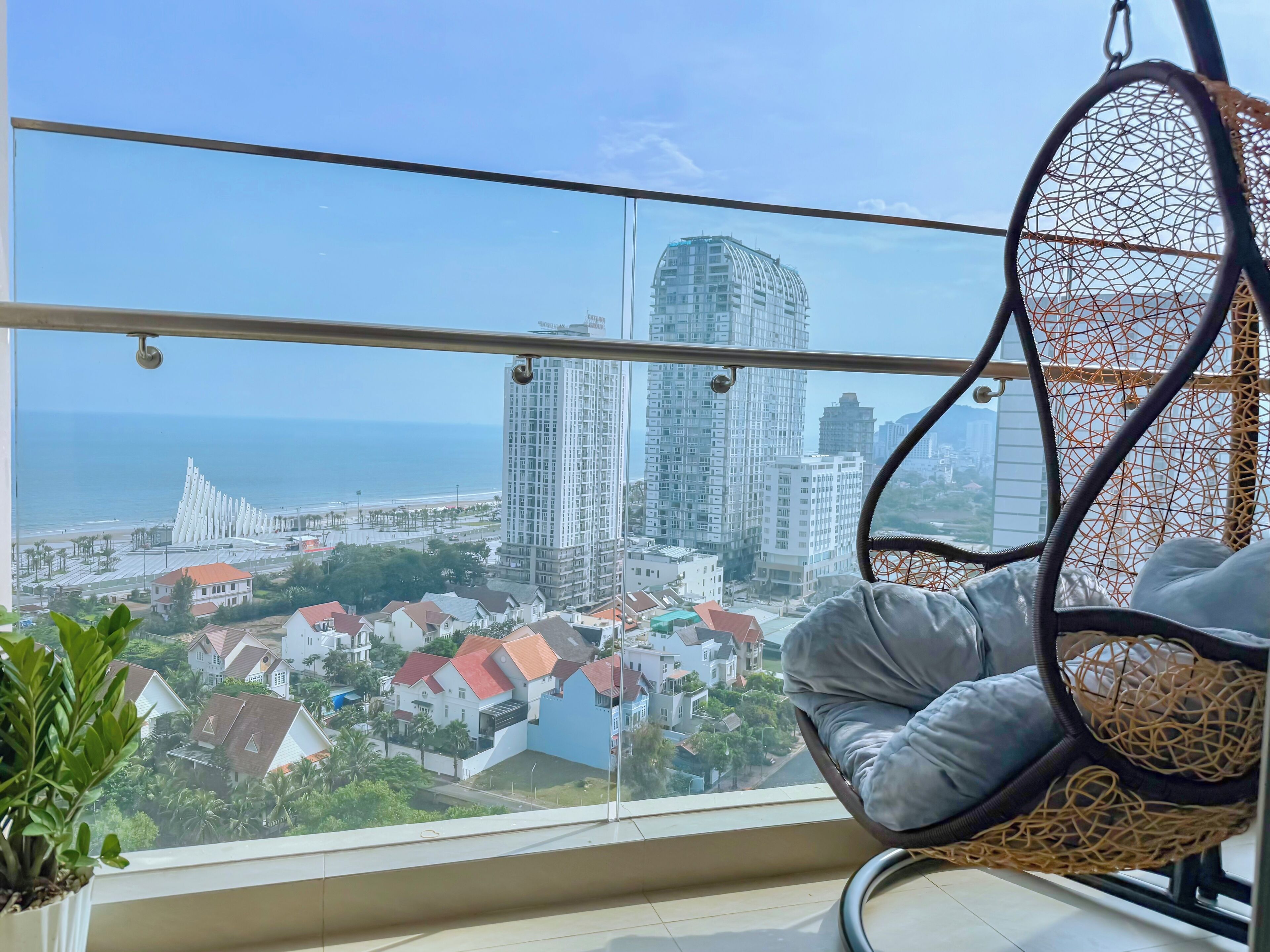 A balcony with a hanging chair, a potted plant, and a view of a cityscape with buildings and a beach.