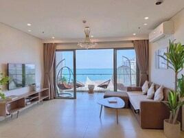 A modern living room with a large TV, comfortable seating, a coffee table, and a view of the sea.