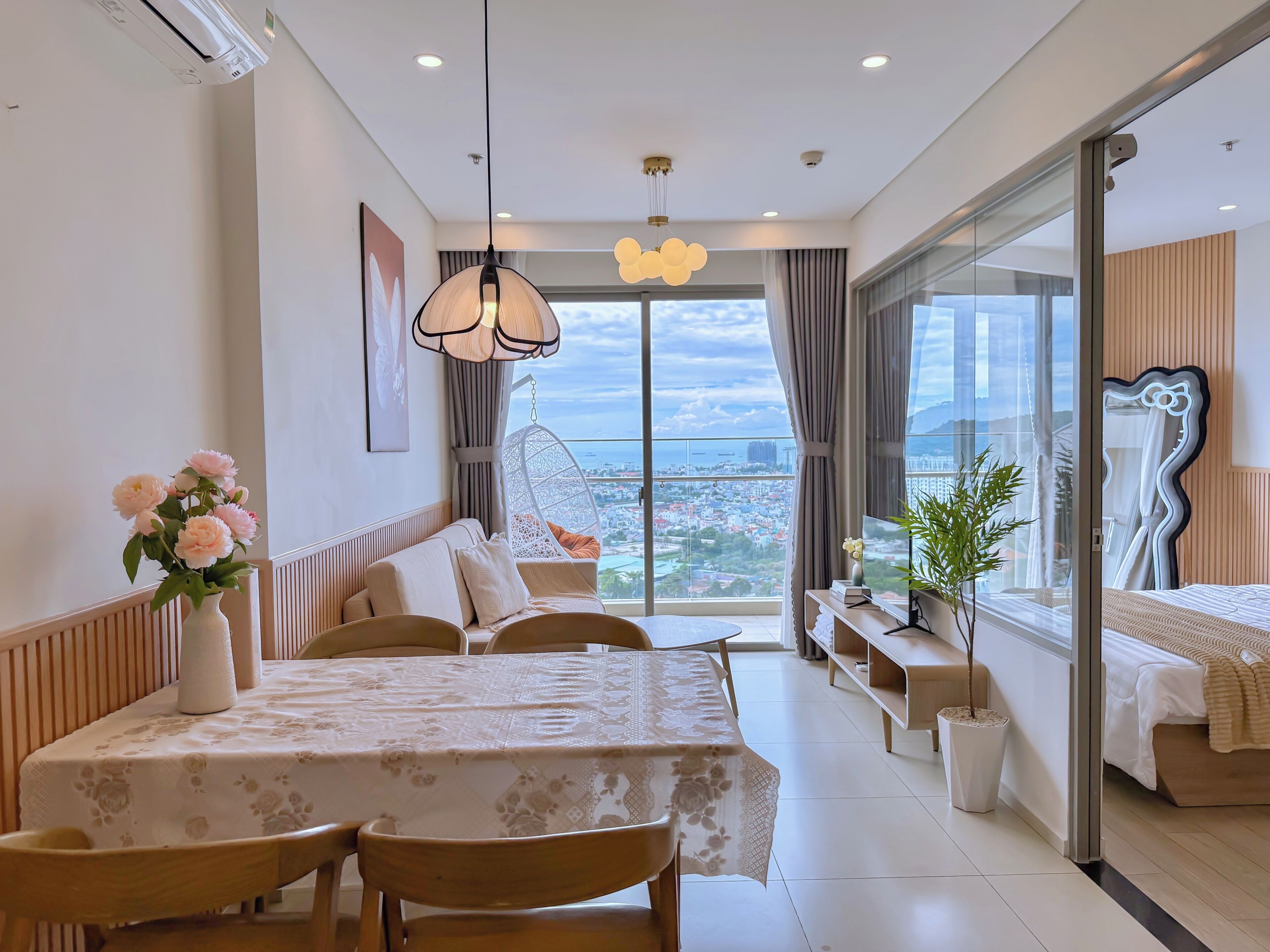 A modern hotel room with a dining area, a hanging chair, a bed, and a large window offering a city view.