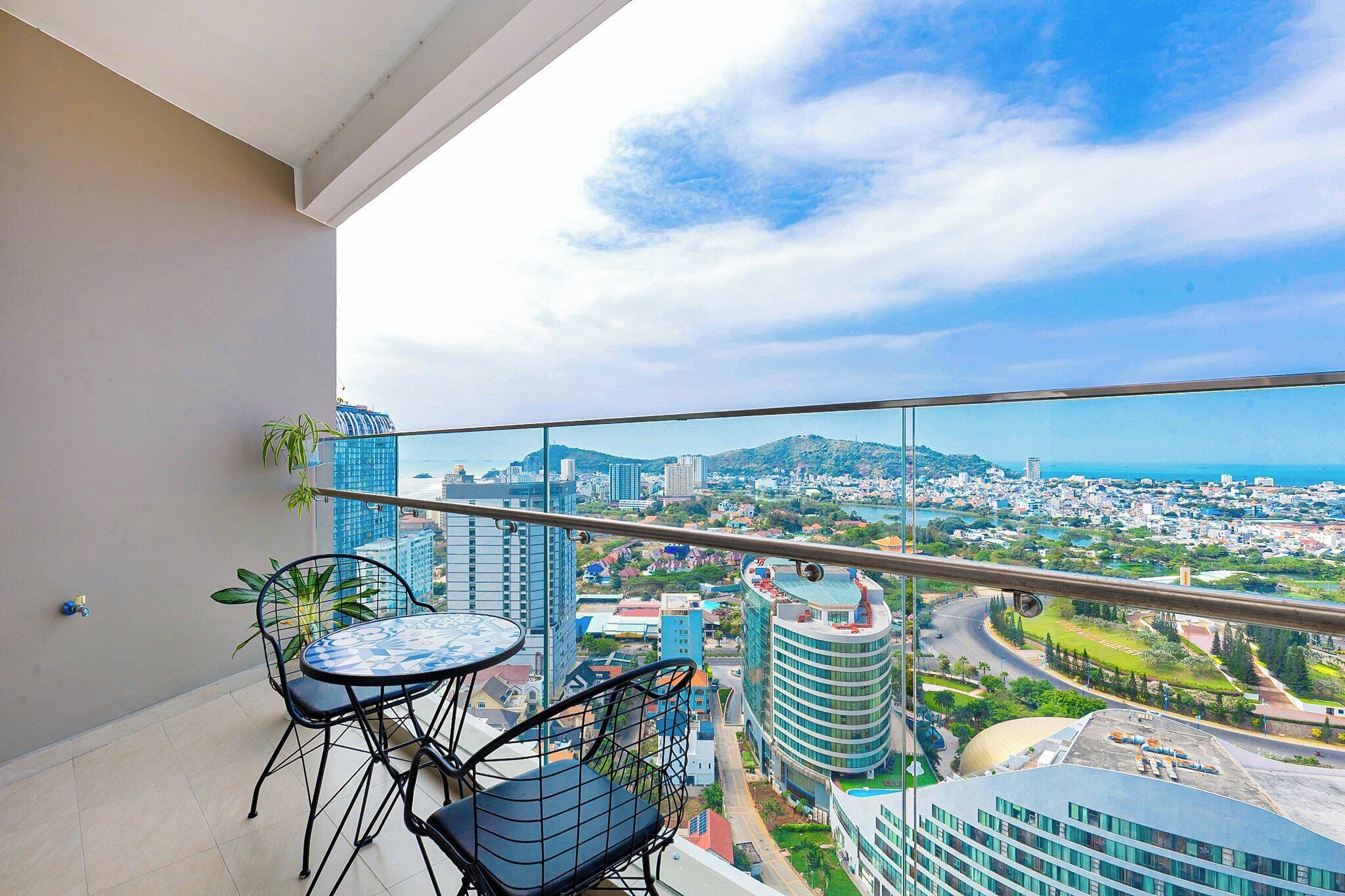 A balcony with a table and chairs, overlooking a cityscape with buildings and a distant view of the sea.