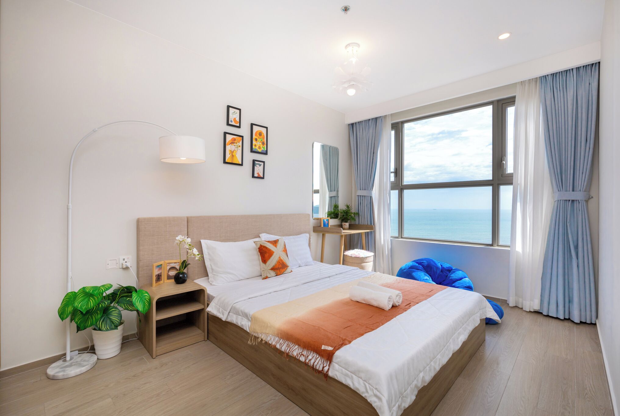 Habitación de hotel moderna con una cama grande, una mesita de noche con una planta, una lámpara de pie y vista al mar a través de la ventana.