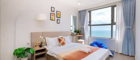 Habitación de hotel moderna con una cama grande, una mesita de noche con una planta, una lámpara de pie y vista al mar a través de la ventana.