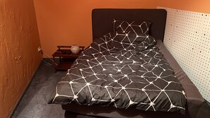 Un lit simple avec une couvre-lit à motifs géométriques, une table de chevet ornée d’un vase et un mur agrémenté d’un panneau perforé.