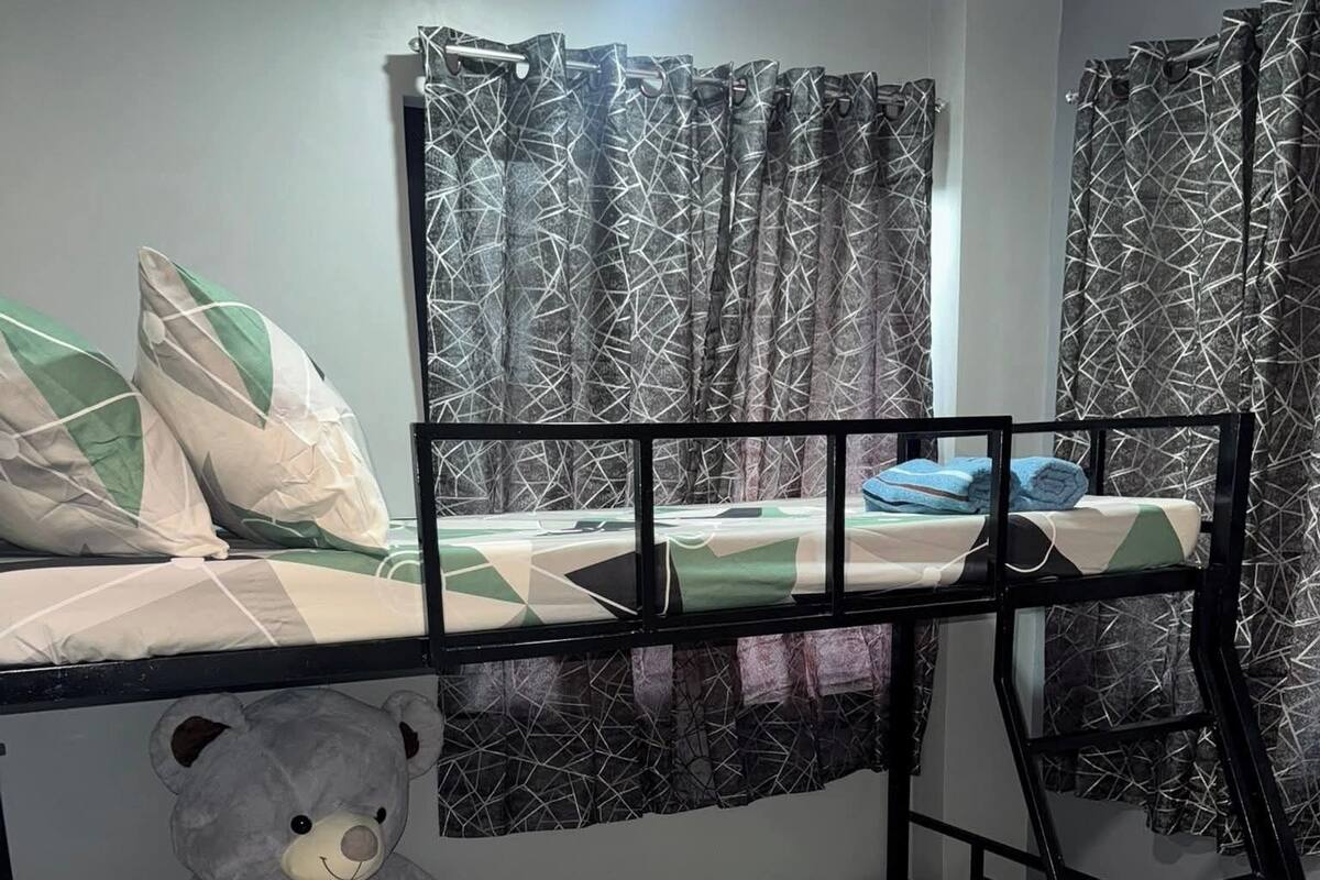 Quarto com beliches, roupa de cama com padrão geométrico, um urso de pelúcia e cortinas com design cruzado.