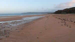 Une vaste plage de sable fin, une mer calme, des affleurements rocheux et une colline qui se dessine à l’horizon.