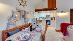 Un coin repas avec une table dressée pour deux personnes, un mur en pierre, une décoration représentant un gouvernail de bateau et, au fond, une cuisine.