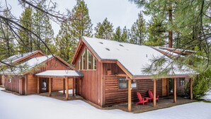 Una cabaña de troncos con techo nevado, fachada de madera y un porche cubierto con sillas rojas.