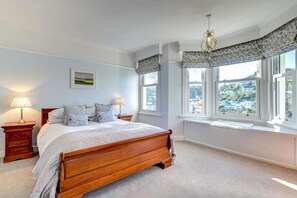 A bedroom with a wooden bed, two bedside tables with lamps, a window with a view of a town, and a chandelier. - 4 Bed in Looe (oc-l30049) (Looe)