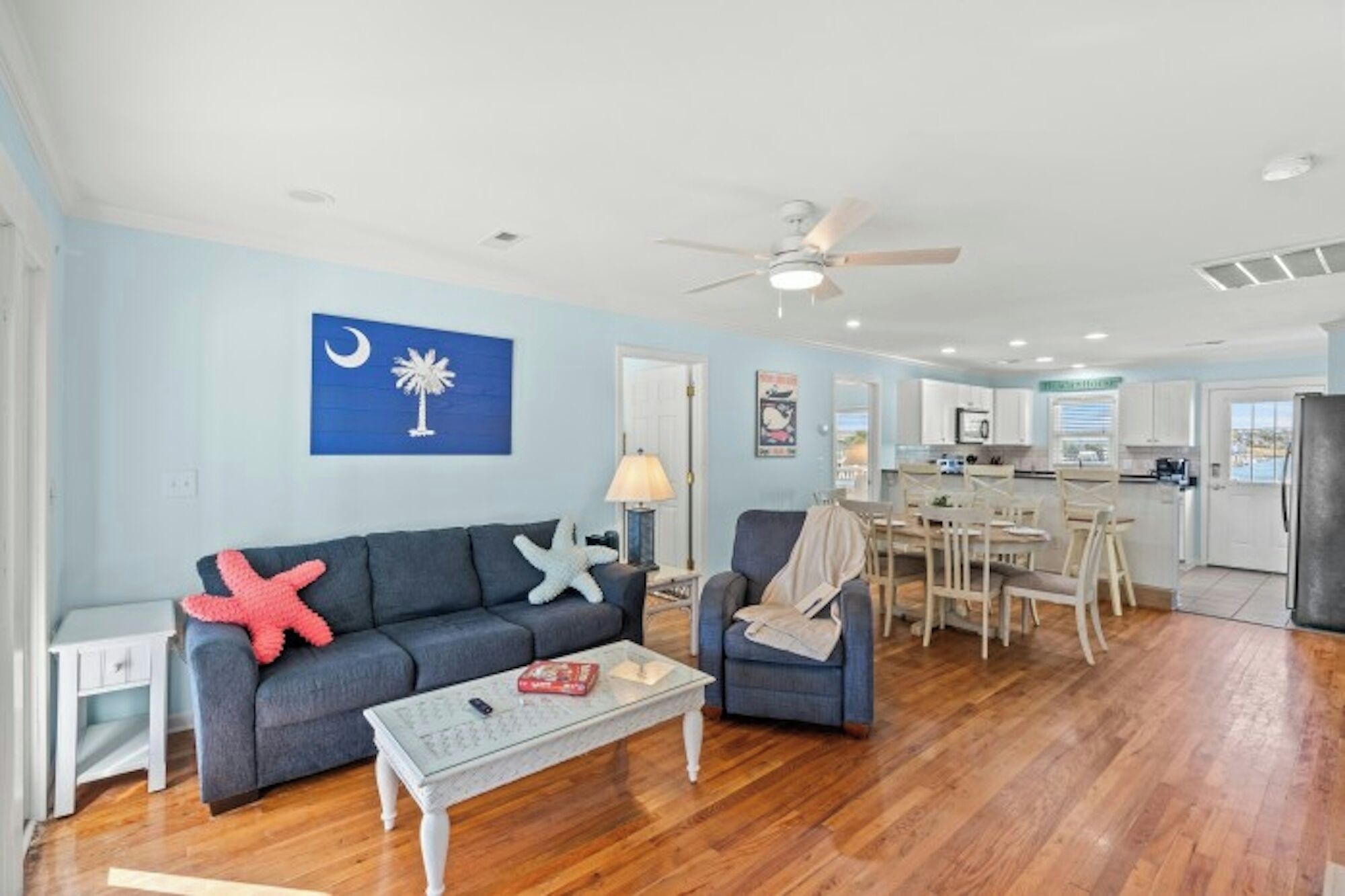 A living room with a sofa, armchairs, a coffee table, and a dining area with a table and chairs. There is a ceiling fan, a kitchen in the background, and a wall with a flag.