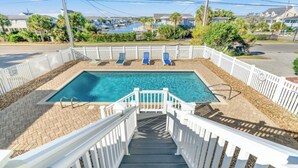 A rectangular swimming pool with a white deck and a white fence surrounding it. There are steps leading up to the pool, and a few lounge chairs are visible in the background. - Shore Beats The Heat- 4BR in Cherry Grove- Pool (North Myrtle Beach)