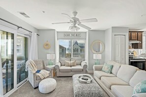 Sala de estar con ventilador de techo, un sofá, una silla y una mesita de centro. Se ve una ventana con persianas y una vista de una casa al exterior.