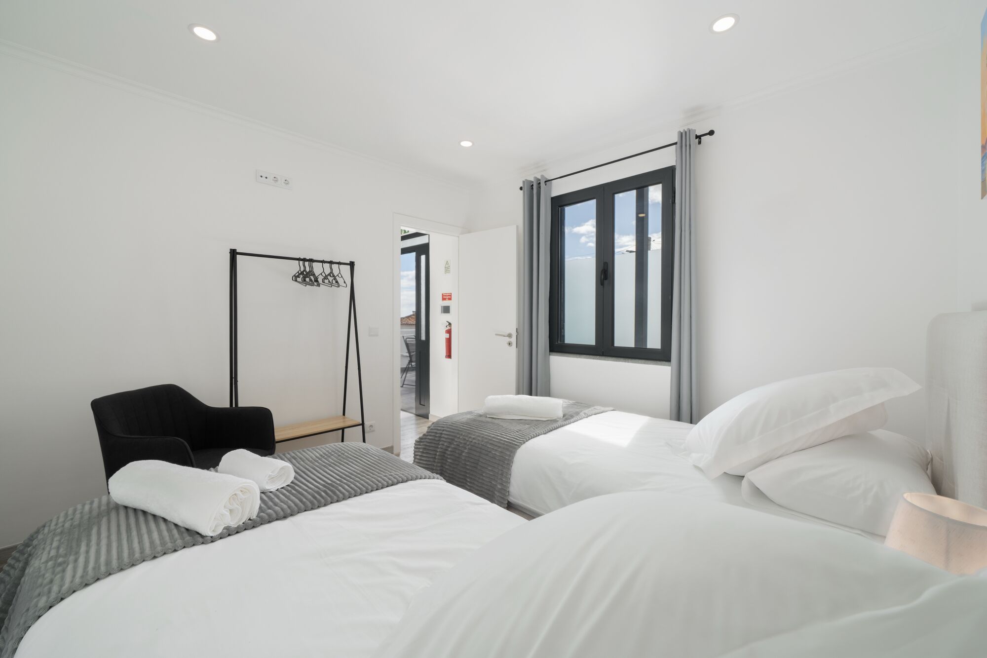 A modern hotel room with two beds, a chair, a coat rack, and a window with curtains.