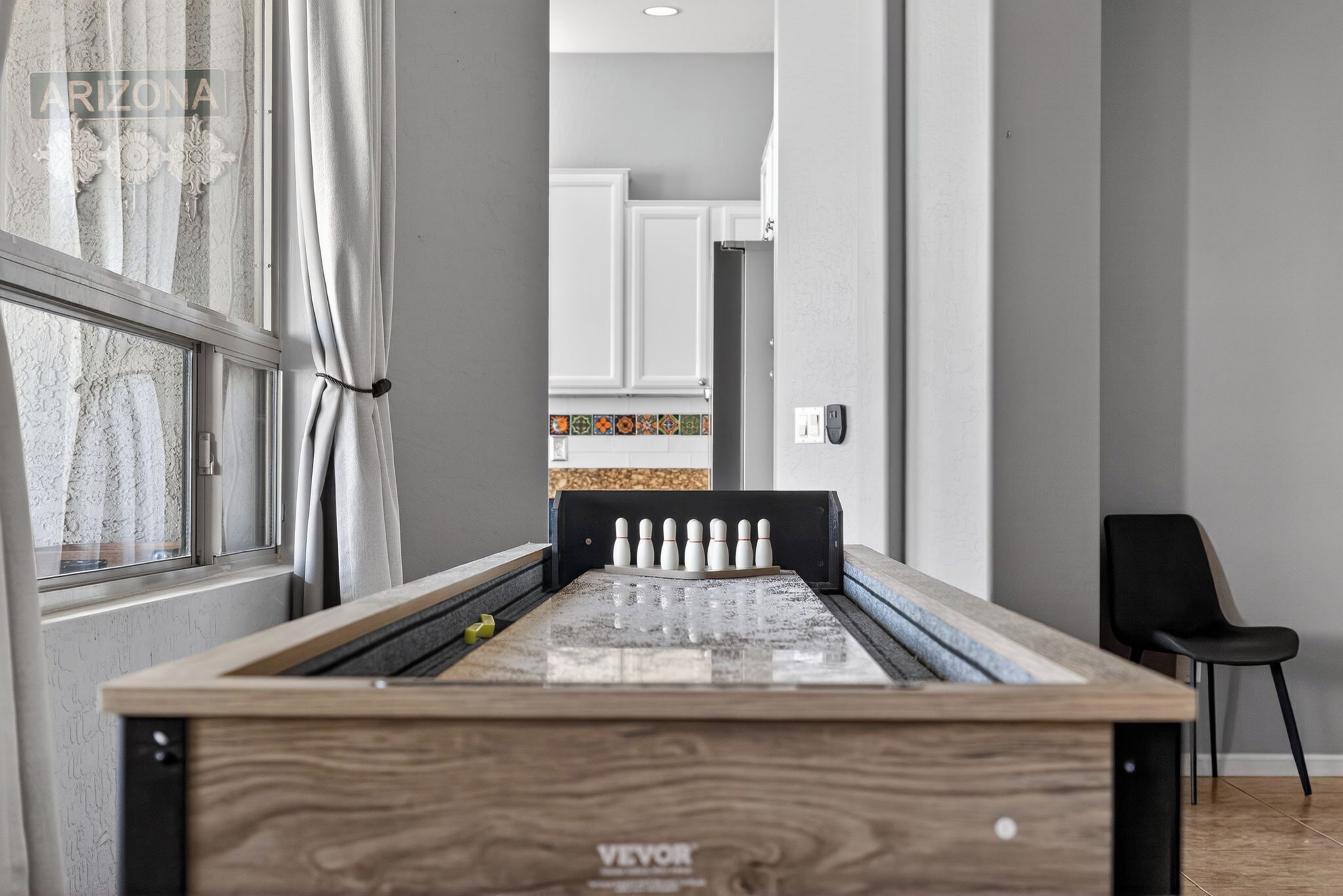 A billiards table with a wooden finish and the brand name 'VEVOR' visible. There is a window with curtains on the left, a black chair to the right, and a kitchen area in the background.